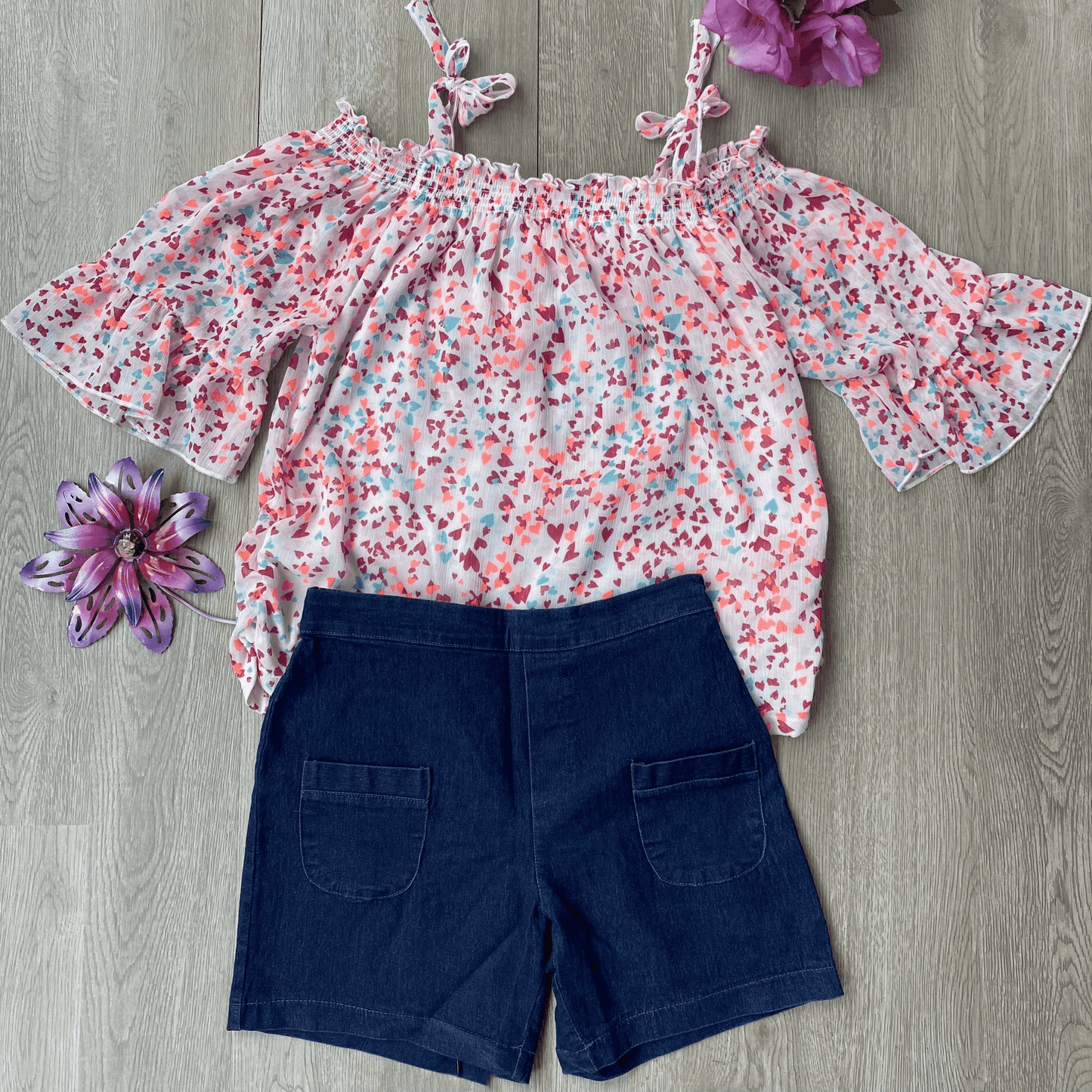 Be summer ready with this cute 2 piece short set featuring an off the shoulder, light weight blouse. The blouse features a pink patterned top with ties on the shoulder. The shorts are denim colored with an elastic waist and 2 pockets on the front.