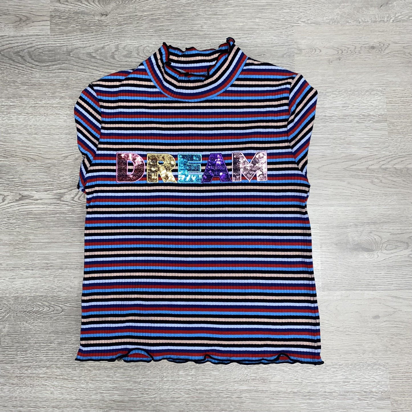 Our ruffle sleeved "DREAM" shirt is a perfect addition to the wardrobe of the little lady in your life. This shirt features long sleeves and a mock turtle neck with scalloped edges. This shirt has red, navy, and blue stripes with the word "DREAM" in sequins.