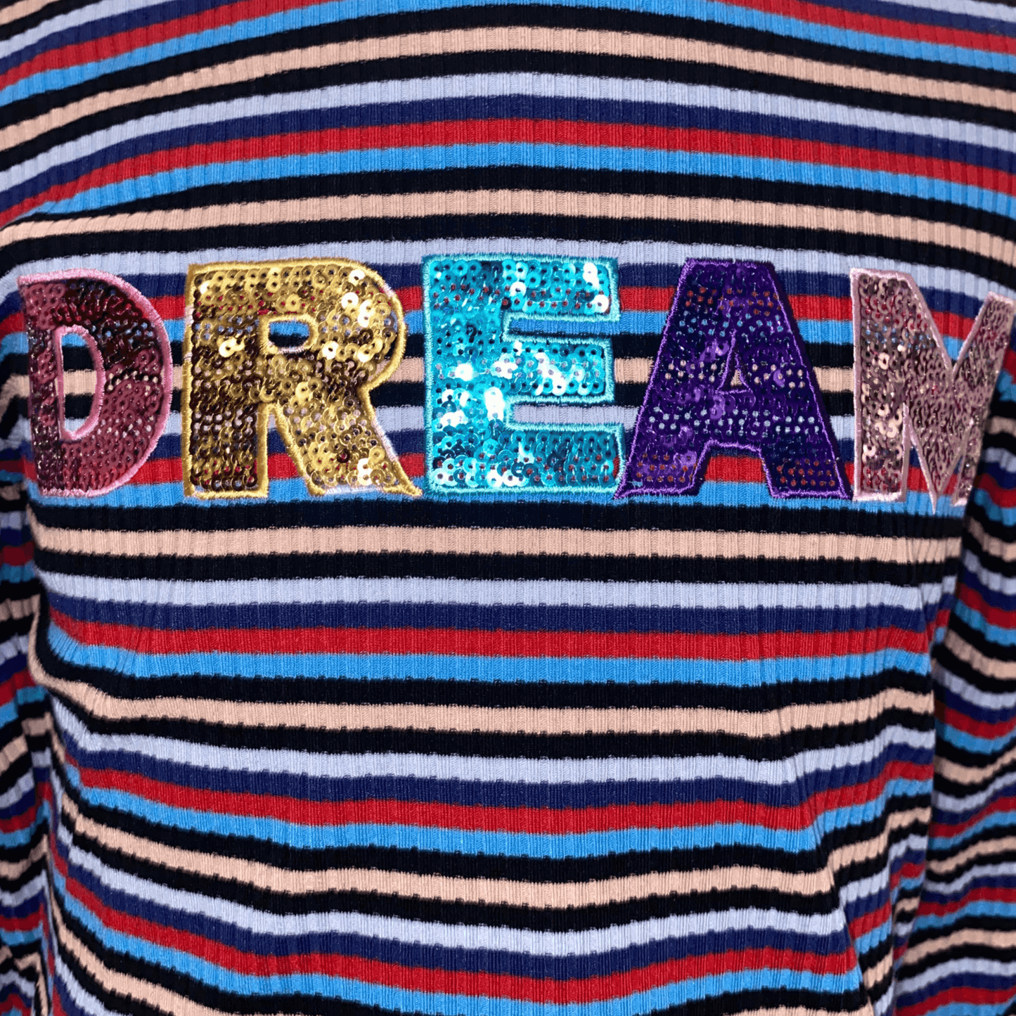Our ruffle sleeved "DREAM" shirt is a perfect addition to the wardrobe of the little lady in your life. This shirt features long sleeves and a mock turtle neck with scalloped edges. This shirt has red, navy, and blue stripes with the word "DREAM" in sequins.